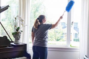 A person stands in a bright room dusting a window with a blue duster. Cleaners Vancouver is featured as a grand piano with sheet music and a red candle sits on the left, while a wooden desk with plants is on the right.