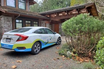 A white and blue sedan with “Get Eco Clean” branding, featuring cleaners Vancouver, is parked in the driveway of a house with a wooden and stone exterior, surrounded by trees, shrubs, and scattered fallen leaves.