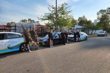 Six people dressed in black stand and lean against three parked cars on a sunny street, with trees, a metal fence, and buildings in the background. Two cars feature a blue and green cleaners Vancouver logo and contact information.