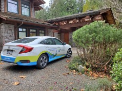 A white and blue RideCo branded car is parked on a gravel driveway in front of a house with stone walls and wooden beams, surrounded by greenery and trees—ideal inspiration for cleaners Vancouver seeking curb appeal.