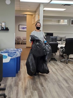 A woman wearing a mask carries two large black garbage bags inside an office with desks, chairs, and recycling bins. Cleaners Vancouver services keep workspaces tidy and organized.