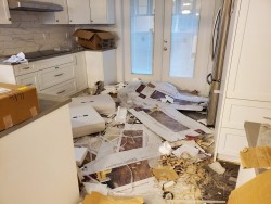 A kitchen with white cabinets is cluttered with construction debris, cardboard boxes, and packing materials scattered across the floor. The space looks untidy and in need of professional attention from cleaners Vancouver.