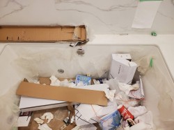A bathtub filled with cardboard, packaging materials, tissue paper, and various trash items looks messy. Plastic sheeting is draped over the tub against marble-patterned walls—an ideal job for cleaners Vancouver residents trust.