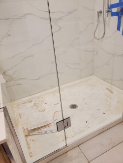 A glass shower enclosure with marble-patterned tiles and a handheld showerhead. The dirty shower floor, scattered with debris, screws, and tape, suggests ongoing renovation—perfect for cleaners Vancouver to restore to pristine condition.