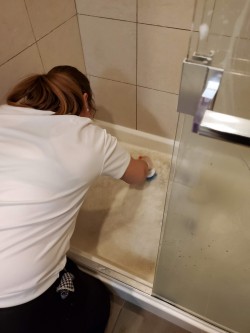 A person in a white shirt is scrubbing the floor of a shower with a sponge, cleaning the dirty surface. The shower has a glass door and tiled walls, showcasing the quality work of cleaners Vancouver offers.