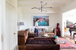 A woman vacuums the floor in a stylish living room with brown leather sofas, a large wall painting, a red guitar case, and a grand piano in the background—showcasing the expertise of cleaners Vancouver clients trust.