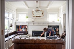 A woman is cleaning a table in a bright living room with a brown leather sofa, built-in bookshelves, a fireplace, and a large mirror above the mantel. Natural light streams in as she uses trusted cleaners Vancouver relies on.